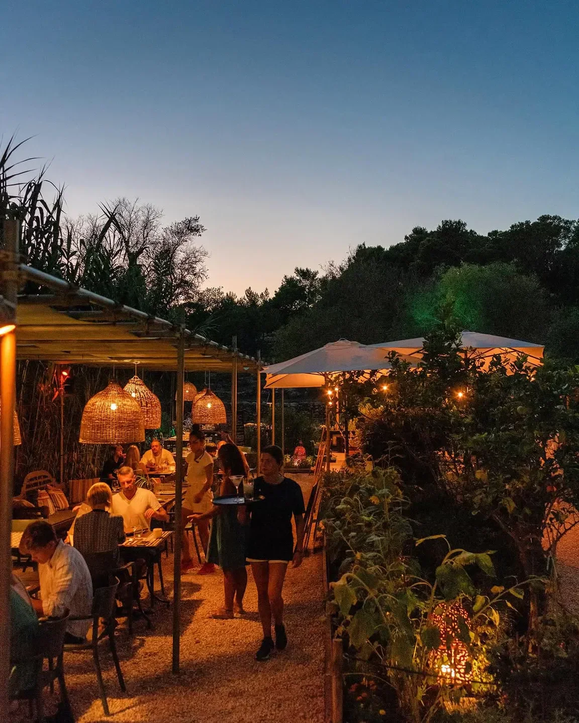 Café Giri in Ibiza: Outdoor dining with wicker lamps and waiter serving drinks at dusk.