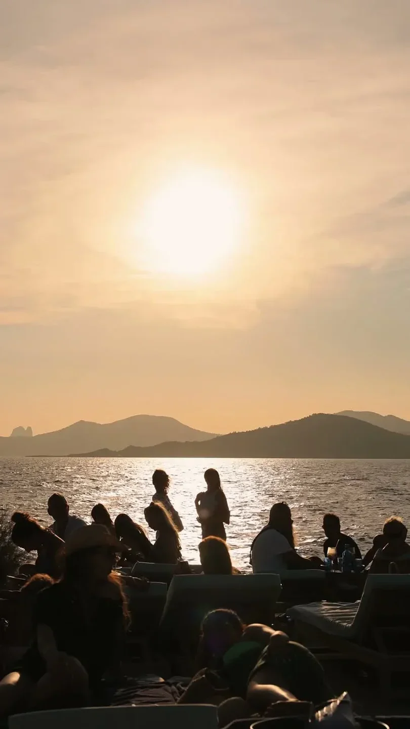 Silhouetted people relax on lounge chairs at Experimental Beach, Ibiza during a golden sunset over the water.